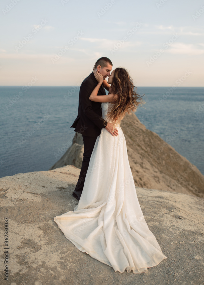 Sensual and beautiful wedding couple passionately embraces on top of the cliff overlooking the sea. A bride in a fashion wedding dress with a chic hem snuggles up to her beloved. Sensual and beautiful wedding couple passionately embraces on top of the cliff overlooking the sea. A bride in a fashion wedding dress with a chic hem snuggles up to her beloved.