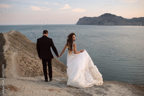 A loving bride and groom walk along the coast of the bay with a view of the cliffs and the ocean. The bride holds a luxurious flowing hemline and turns smiling at the camera. photo