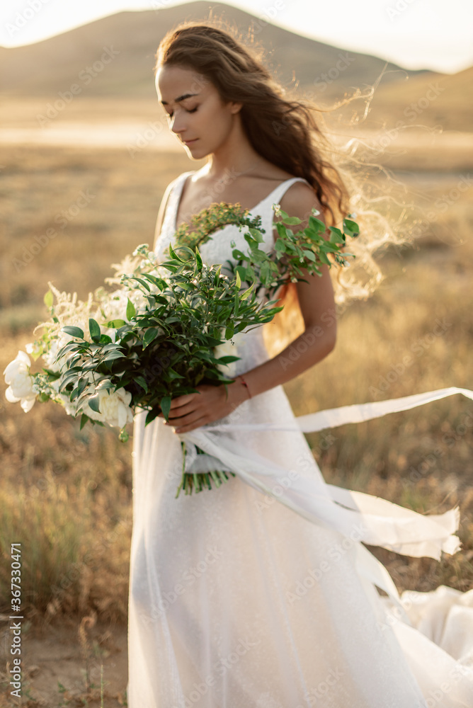 Beautiful and tender bride with wavy golden hair in fashionable wedding dress with luxury wedding peony bouquet stands in the sunset light against mountains. Beautiful and tender bride with wavy golden hair in fashionable wedding dress with luxury wedding peony bouquet stands in the sunset light against mountains.
