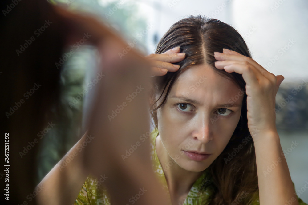 Portrait of a beautiful young woman examining her scalp and hair in front of the mirror, hair roots, color, grey hair, hair loss or dry scalp problem Portrait of a beautiful young woman examining her scalp and hair in front of the mirror, hair roots, color, grey hair, hair loss or dry scalp problem