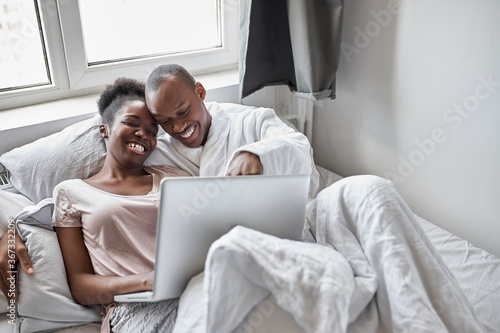 black afroamerican woman lie on bed with laptop, couple in domestic wear lie on bed together, woman works from home