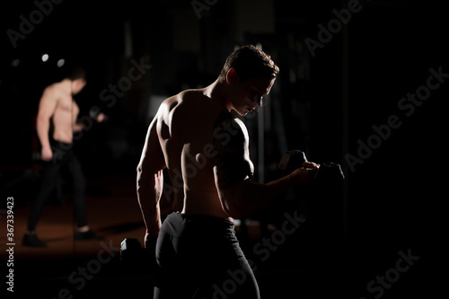 Handsome power athletic guy bodybuilder doing exercises with dumbbell. Fitness muscular body on dark background.