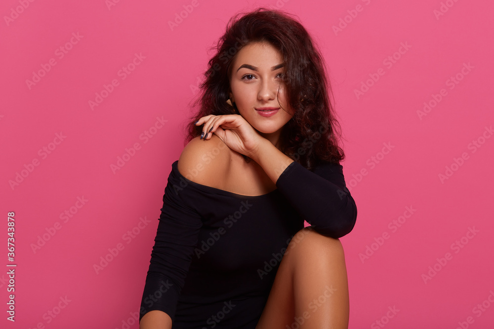 Picture of charming female with pleasant appearance, looking at camera, wearing black bodysuit, posing isolated over pink background, keeping hand near bare shoulders. Picture of charming female with pleasant appearance, looking at camera, wearing black bodysuit, posing isolated over pink background, keeping hand near bare shoulders.