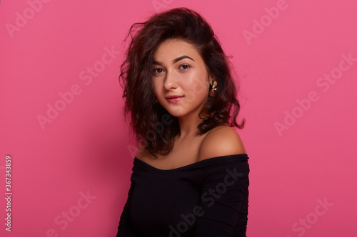 Brunette female posing with bare shoulders against pink wall, having dark wavy hair, looking at camera, looks gentle and romantic, wearing black shirt.