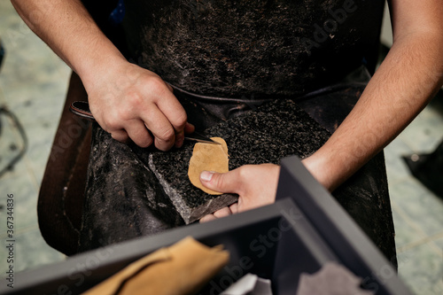 Male cobler sitting at his vintage looking workshop and cutting leather fabric for shoe repairing photo