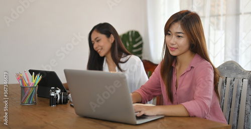 Women are using a computer tablet and laptop while sitting together at the wooden table.