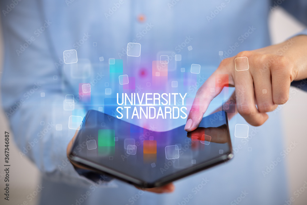 Young man holding a foldable smartphone with UNIVERSITY STANDARDS inscription, educational concept Young man holding a foldable smartphone with UNIVERSITY STANDARDS inscription, educational concept