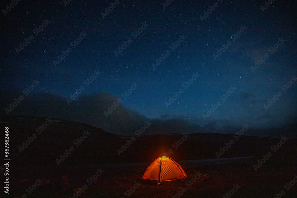 The concept of outdoor recreation. Glowing orange tent in the mountains under dramatic evening sky. Red sunset and mountains in the background. Summer landscape.