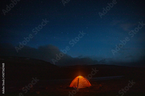 The concept of outdoor recreation. Glowing orange tent in the mountains under dramatic evening sky. Red sunset and mountains in the background. Summer landscape.