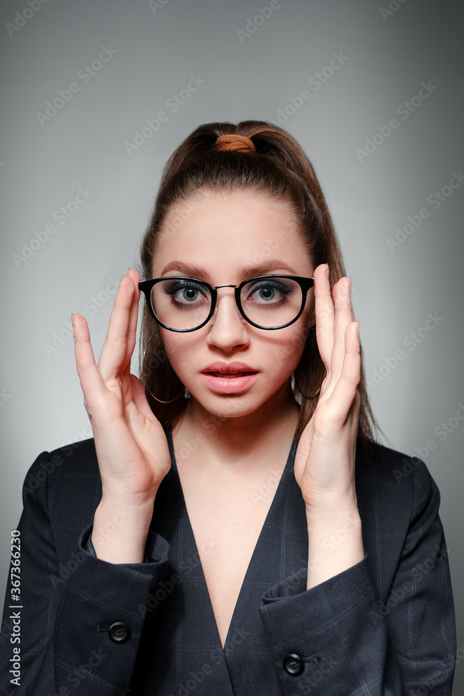 A young girl 20-25 years old in glasses, a jacket and with a tail in the image of a teacher poses on a gray background and shows different emotions A young girl 20-25 years old in glasses, a jacket and with a tail in the image of a teacher poses on a gray background and shows different emotions