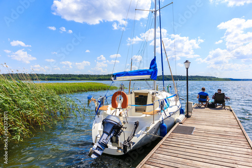 Two unidentified men fishing on pier and sailing boat mooring on lake shore in Krzyze village port on sunny summer day, Mazury Lake District, Poland