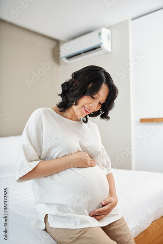 Smiling beautiful young Vietnamese woman touching her belly and enjoying her pregnancy
