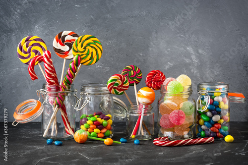 Colored lollipops, colorful round candies and marmalade in glass jars on a black stone table with a gray backdrop. Selective focus. Festive background with sweets.