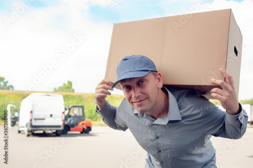 A man in a gray suit in his hands with a cardboard box against the background of the van. Concept on the topic of postal delivery. © Sergey