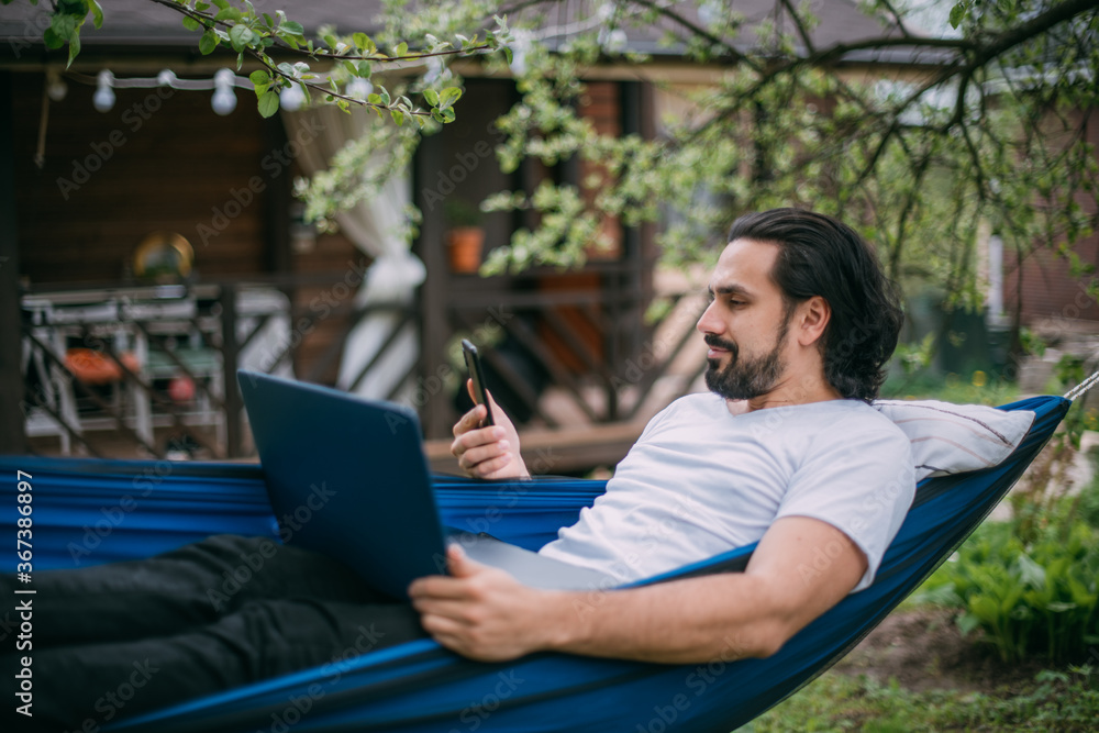 A man works with a laptop and a phone in a hammock in a country house A man works with a laptop and a phone in a hammock in a country house
