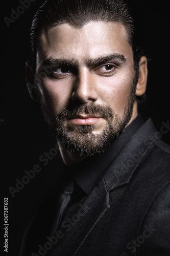 Fotografija Young white Caucasian man on a black background in a black jacket, shirt and black tie looks out from under his forehead
