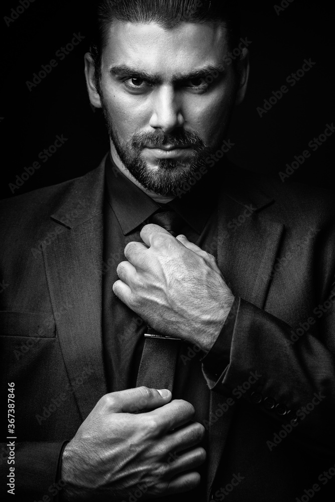Portrait of a white Caucasian male brunet in a black classic suit, black  shirt and black tie over a black background. Fashionable portrait of an  attractive man with dark hair. Stock Photo |