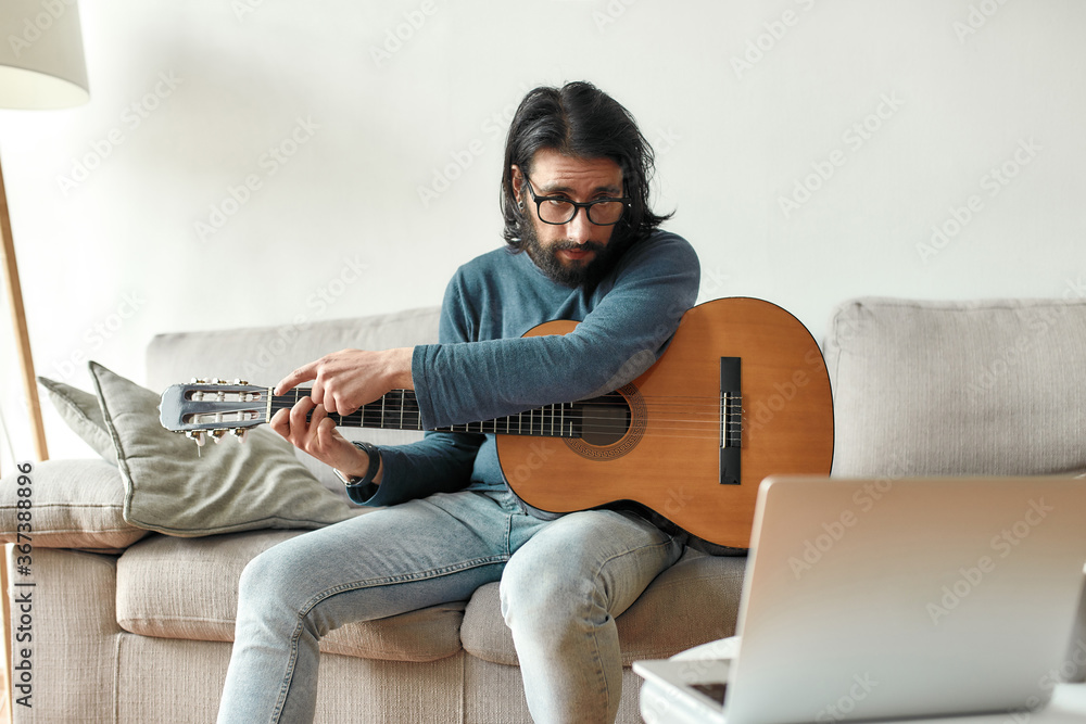 Young caucasian man sitting on sofa at home and adjusting acoustic guitar, watching guitar lesson online on laptop