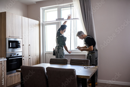 Canvas Print Young professional worker in uniform using tape measure, measuring window for installing blinds, while his aged colleague making notes