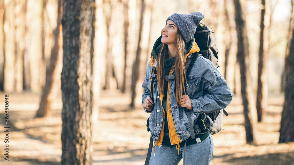 Half-length horizontal portrait of a young attractive tourist girl with a charming smile. Caucasian white girl with a large hiking backpack in the forest. Autumn colors.