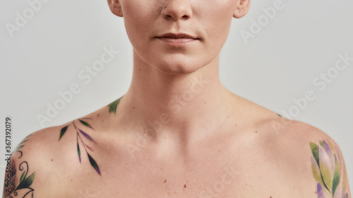 Cropped portrait of a young attractive naked tattooed woman with perfect skin looking at camera isolated over light background