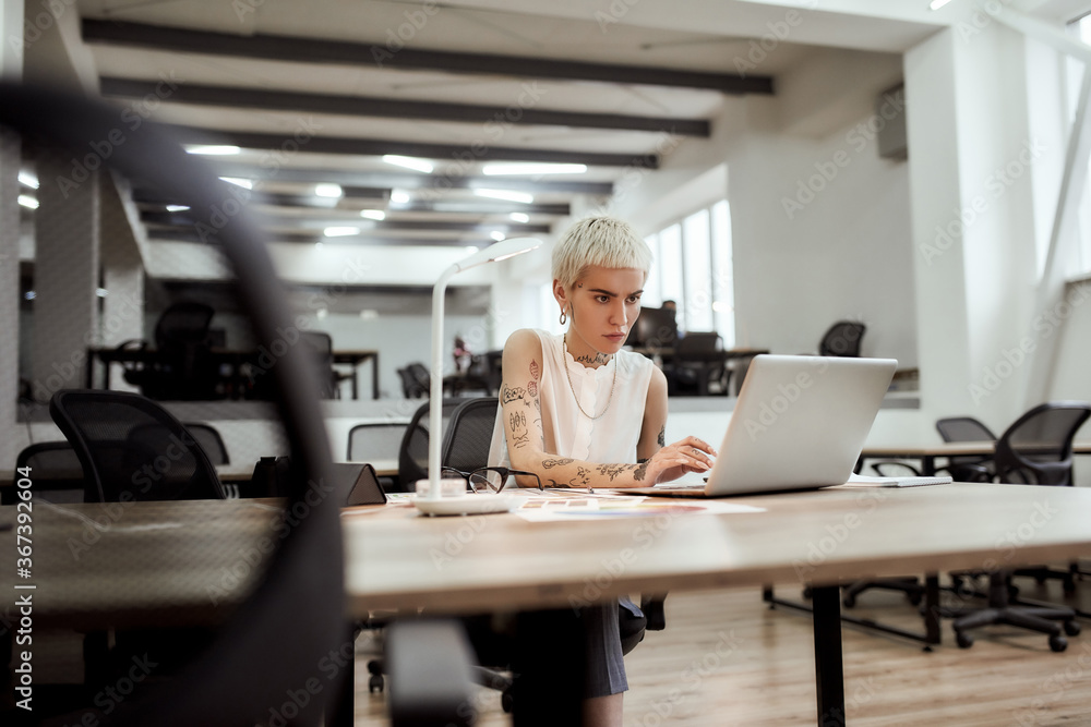 Focused young tattooed female designer using laptop while working at her desk in the modern office. Designer workplace Focused young tattooed female designer using laptop while working at her desk in the modern office. Designer workplace