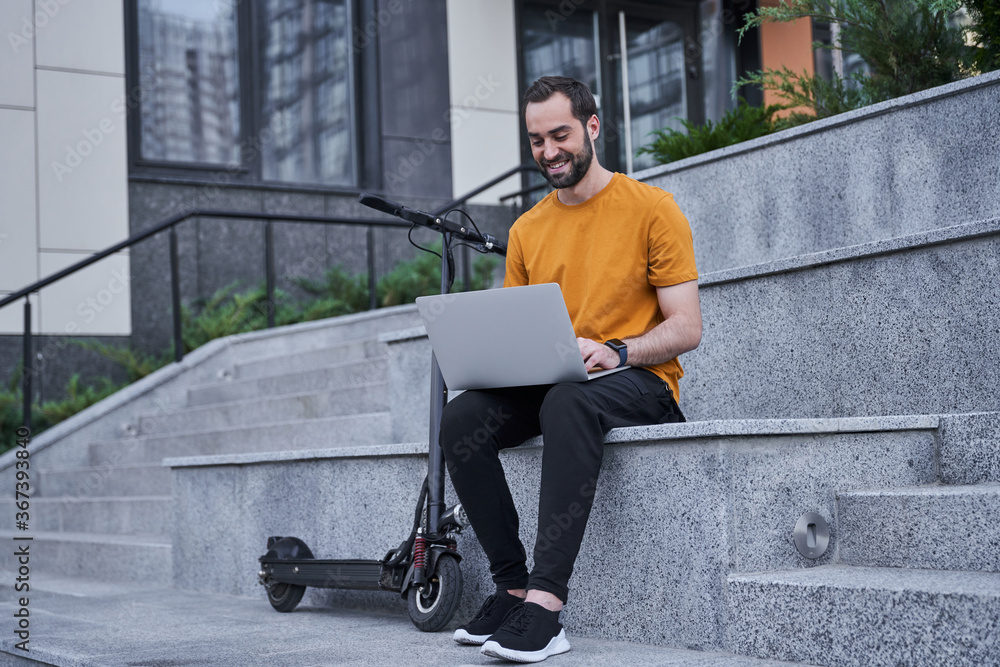 Jolly young man with scooter using laptop outdoors Jolly young man with scooter using laptop outdoors