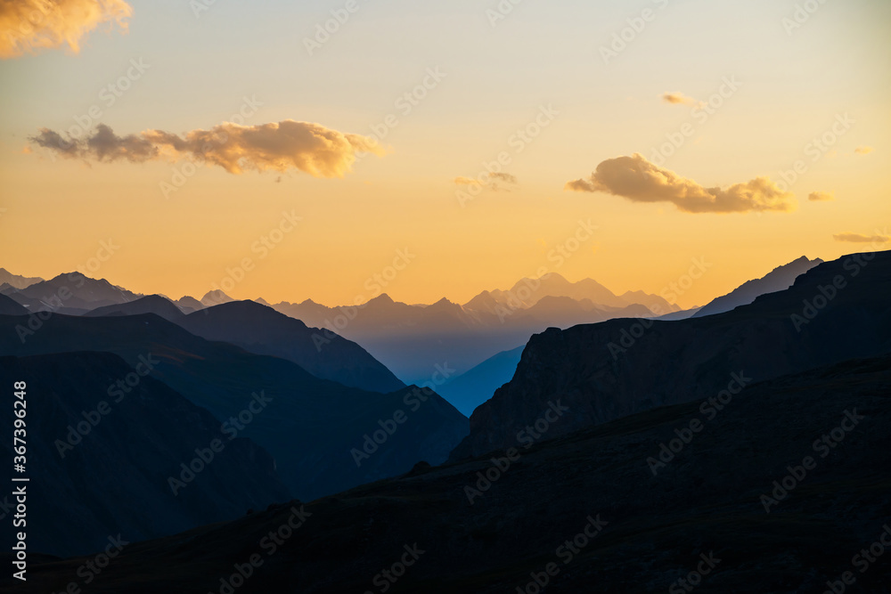 Colorful dawn landscape with beautiful blue mountains silhouettes and golden gradient sky with clouds. Vivid mountain scenery with picturesque multicolor sunset. Scenic sunrise view to mountain range.