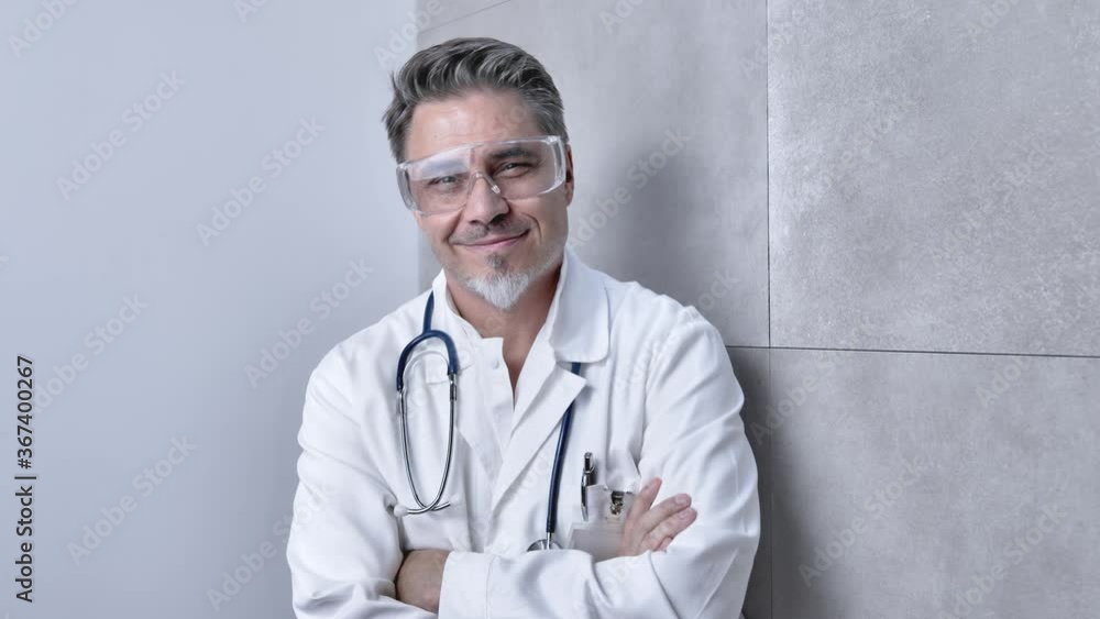Portrait of trustworthy older smart doctor with gray hair wearing safety glasses and white lab coat standing against gray wall, smiling. Copy space. Portrait of trustworthy older smart doctor with gray hair wearing safety glasses and white lab coat standing against gray wall, smiling. Copy space.
