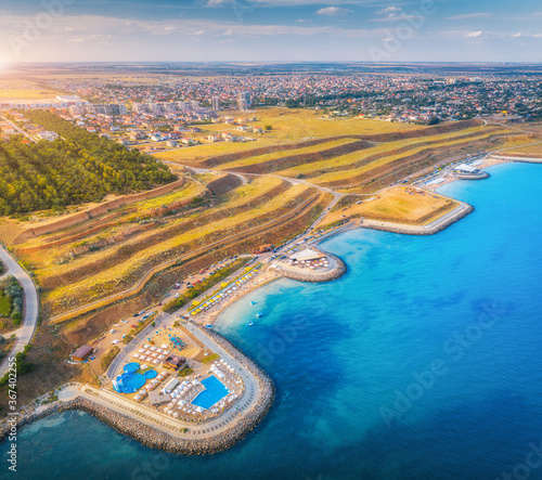 Aerial view of beautiful sandy beach, blue sea, restaurants on the promenade, pool, umbrellas, swimming people in clear water, green trees at sunset in summer. Top view of seafront, yellow hills, city