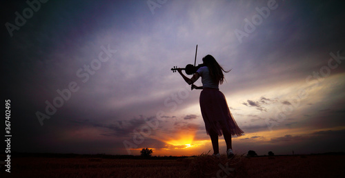Romantic young woman with loose hair playing the violin in a field at sunset
