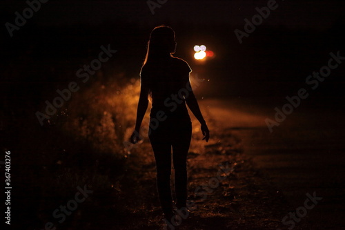 Silhouette of young slender woman in the backlight of car headlights on the road