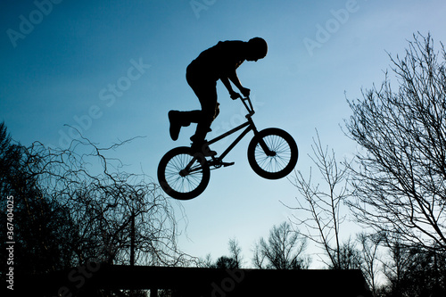 Silhouette of a man jumping on a bicycle against a blue sky with white clouds. Guy holding bike handlebar and doing trick.