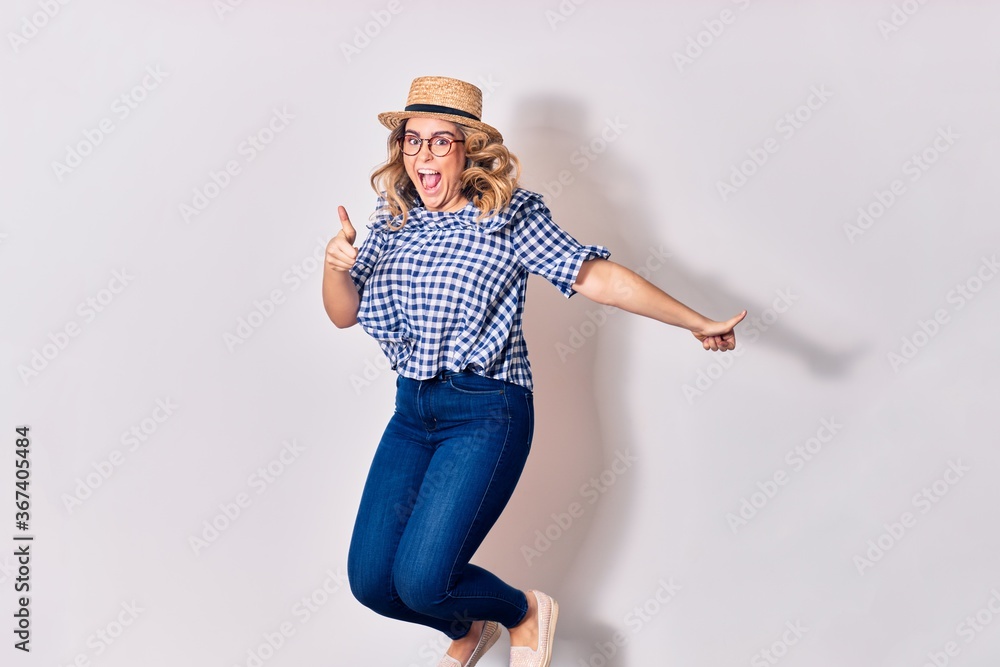 Young beautiful blonde woman wearing glasses and summer hat smiling happy. Jumping with smile on face over isolated white background. Young beautiful blonde woman wearing glasses and summer hat smiling happy. Jumping with smile on face over isolated white background.