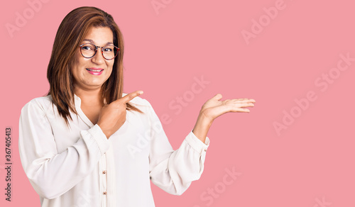 Middle age latin woman wearing casual clothes and glasses amazed and smiling to the camera while presenting with hand and pointing with finger.