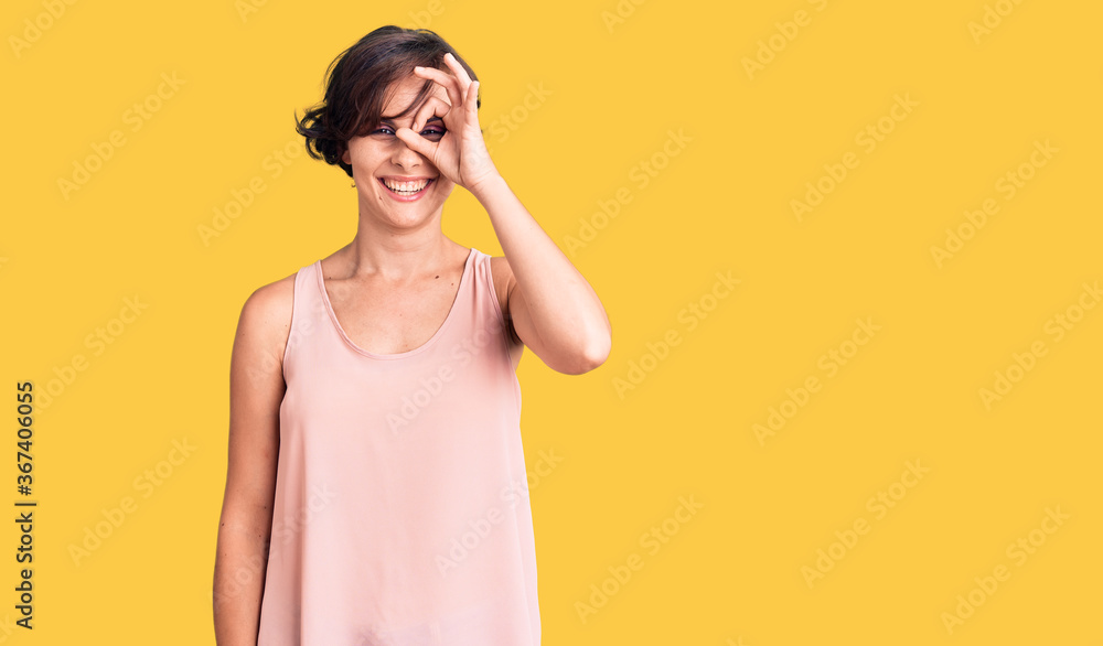 Beautiful young woman with short hair wearing casual style with sleeveless shirt doing ok gesture with hand smiling, eye looking through fingers with happy face. Beautiful young woman with short hair wearing casual style with sleeveless shirt doing ok gesture with hand smiling, eye looking through fingers with happy face.