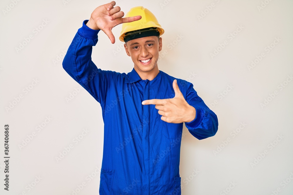 Young hispanic boy wearing worker uniform and hardhat smiling making frame with hands and fingers with happy face. creativity and photography concept.