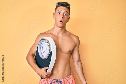 Young hispanic boy shirtless wearing swimwear and holding weighing machine scared and amazed with open mouth for surprise  disbelief face