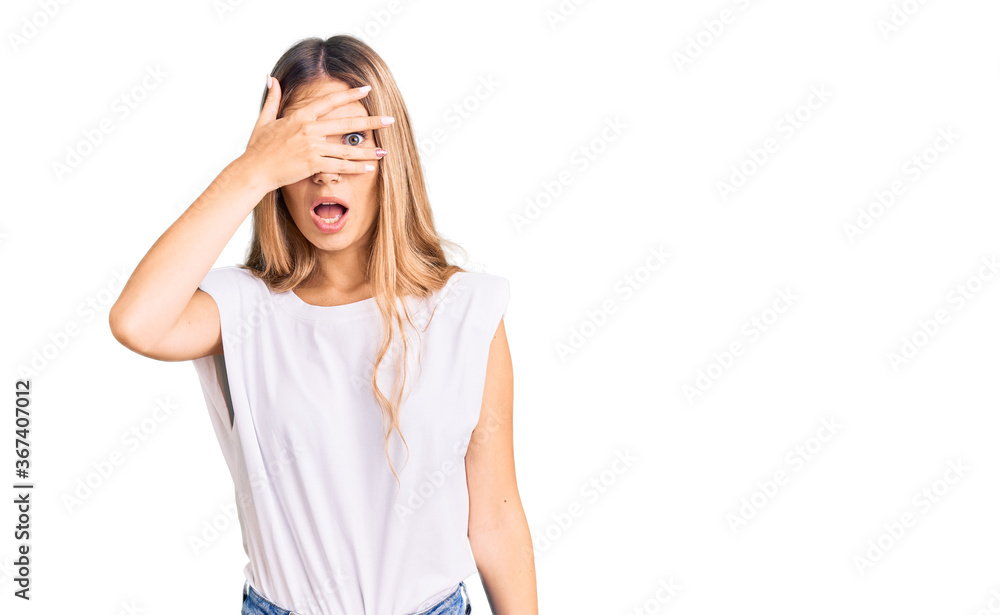 Beautiful caucasian woman with blonde hair wearing casual white tshirt peeking in shock covering face and eyes with hand, looking through fingers with embarrassed expression. Beautiful caucasian woman with blonde hair wearing casual white tshirt peeking in shock covering face and eyes with hand, looking through fingers with embarrassed expression.