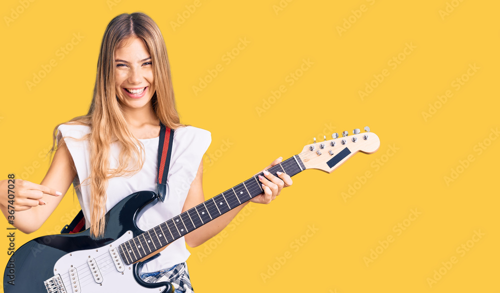Beautiful caucasian woman with blonde hair playing electric guitar smiling happy pointing with hand and finger Beautiful caucasian woman with blonde hair playing electric guitar smiling happy pointing with hand and finger