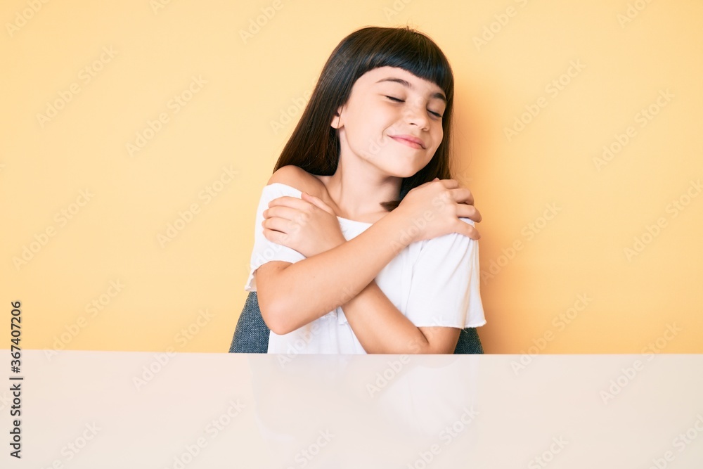 Young little girl with bang wearing casual clothes sitting on the table hugging oneself happy and positive, smiling confident. self love and self care Young little girl with bang wearing casual clothes sitting on the table hugging oneself happy and positive, smiling confident. self love and self care