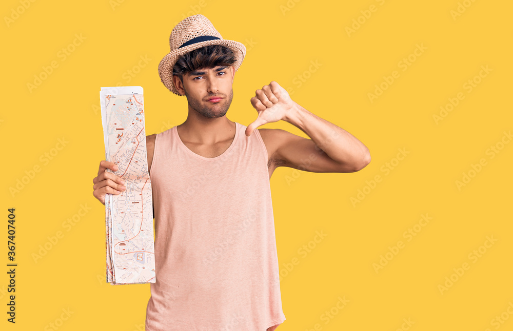 Young hispanic man wearing summer hat holding map with angry face, negative sign showing dislike with thumbs down, rejection concept Young hispanic man wearing summer hat holding map with angry face, negative sign showing dislike with thumbs down, rejection concept
