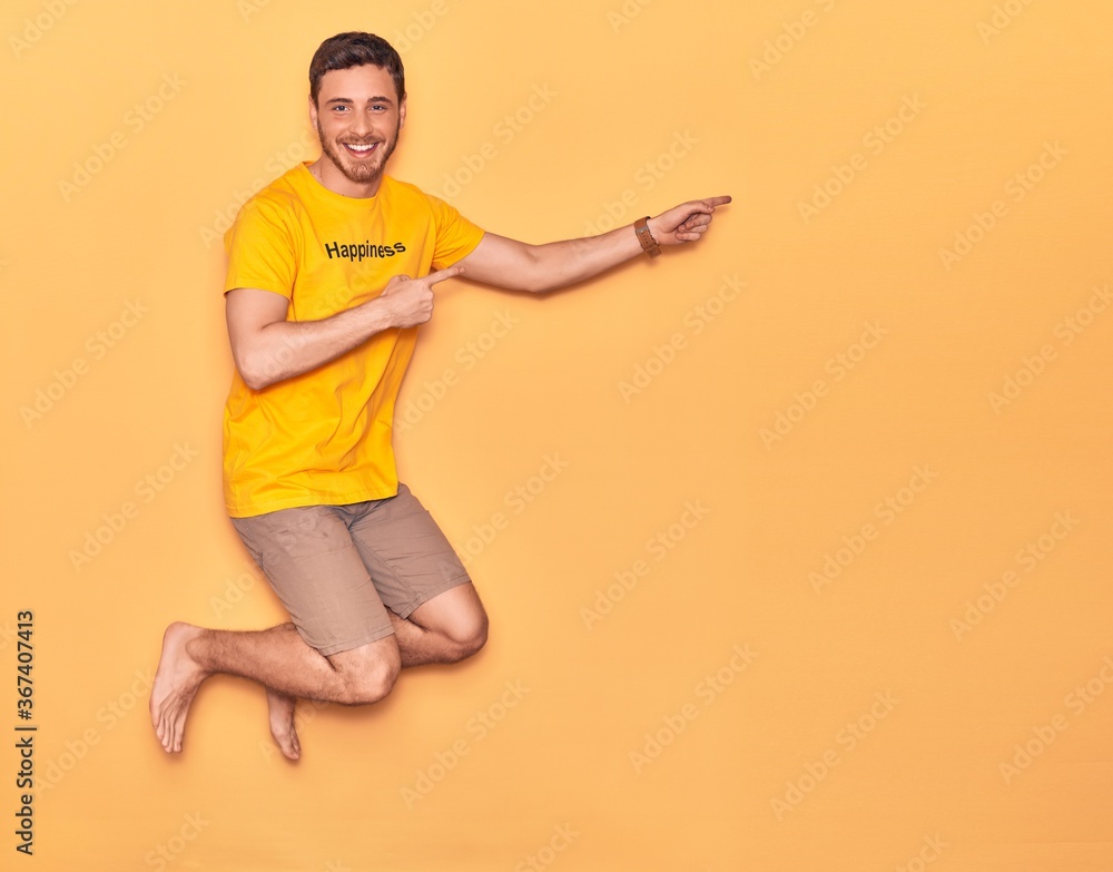 Young handsome hispanic man wearing t shirt with happiness word message smiling happy. Jumping with smile on face over isolated yellow background Young handsome hispanic man wearing t shirt with happiness word message smiling happy. Jumping with smile on face over isolated yellow background