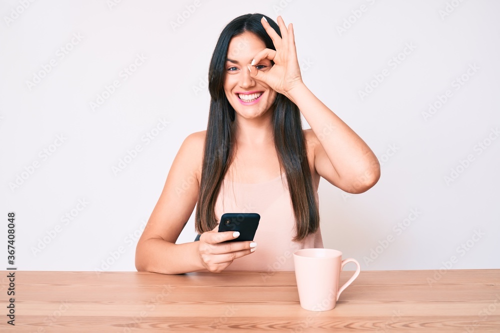 Young caucasian woman sitting at the desk using smartphone drinking coffee smiling happy doing ok sign with hand on eye looking through fingers