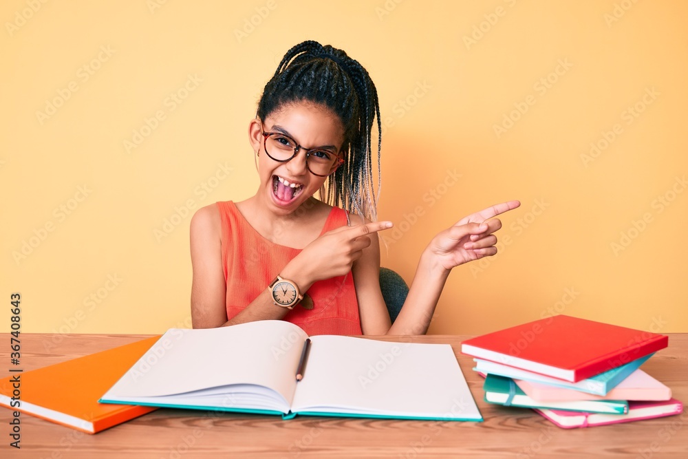 Young african american girl child with braids studying for school exam smiling and looking at the camera pointing with two hands and fingers to the side. Young african american girl child with braids studying for school exam smiling and looking at the camera pointing with two hands and fingers to the side.
