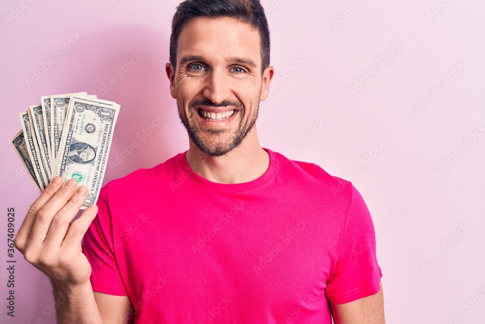Young handsome man holding bunch of dollars banknotes over isolated pink background looking positive and happy standing and smiling with a confident smile showing teeth Young handsome man holding bunch of dollars banknotes over isolated pink background looking positive and happy standing and smiling with a confident smile showing teeth