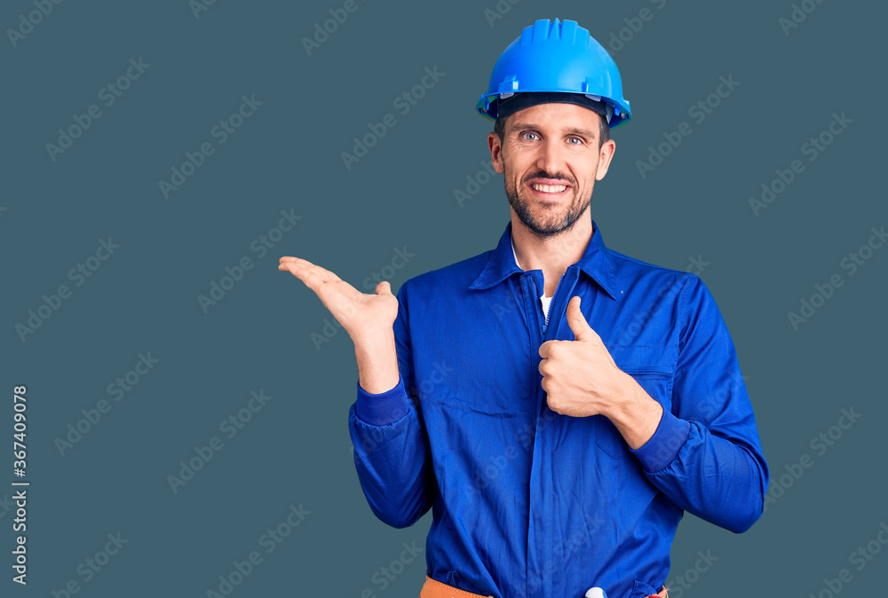 Young handsome man wearing worker uniform and hardhat showing palm hand and doing ok gesture with thumbs up, smiling happy and cheerful Young handsome man wearing worker uniform and hardhat showing palm hand and doing ok gesture with thumbs up, smiling happy and cheerful
