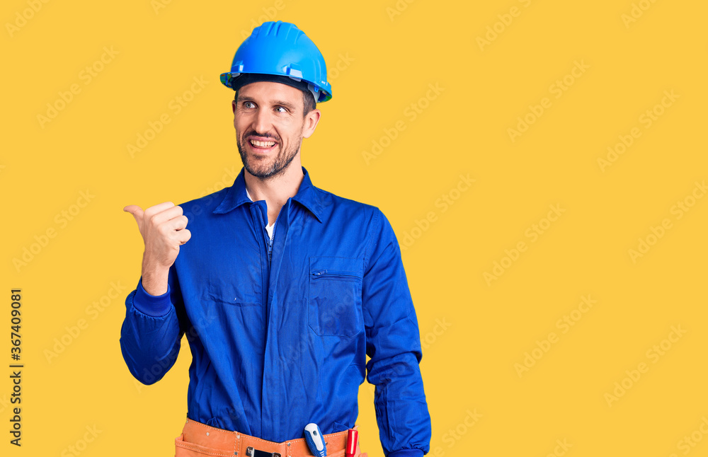 Young handsome man wearing worker uniform and hardhat smiling with happy face looking and pointing to the side with thumb up.
