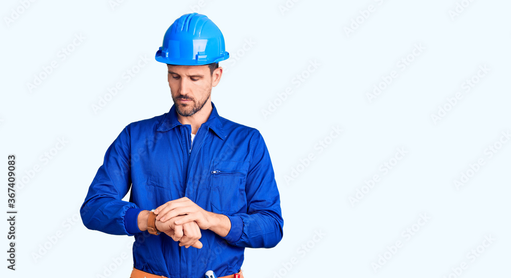 Young handsome man wearing worker uniform and hardhat in hurry pointing to watch time, impatience, upset and angry for deadline delay
