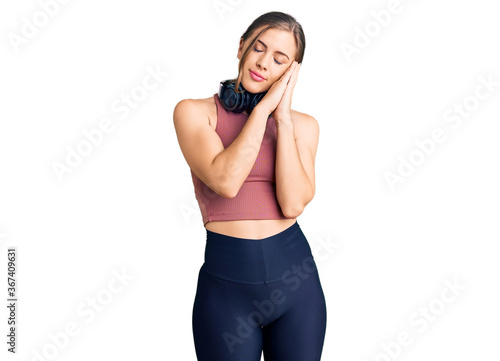 Beautiful caucasian young woman wearing gym clothes and using headphones sleeping tired dreaming and posing with hands together while smiling with closed eyes.